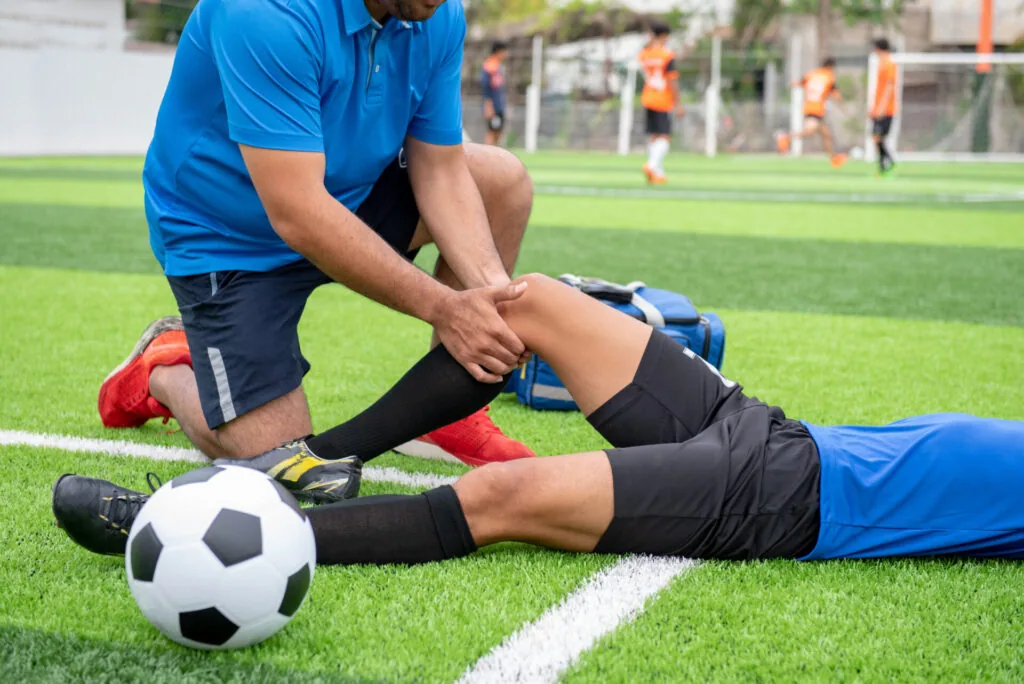 Athletic trainer examining soccer player's leg injury on field while other players practice in background