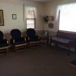Chiropractic office waiting room with glider chairs, wooden bench, ceiling fan, and windows with blinds