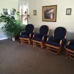 Medical waiting room with wooden glider chairs upholstered in blue and orange fabric, potted plant, framed artwork, and ceiling fan